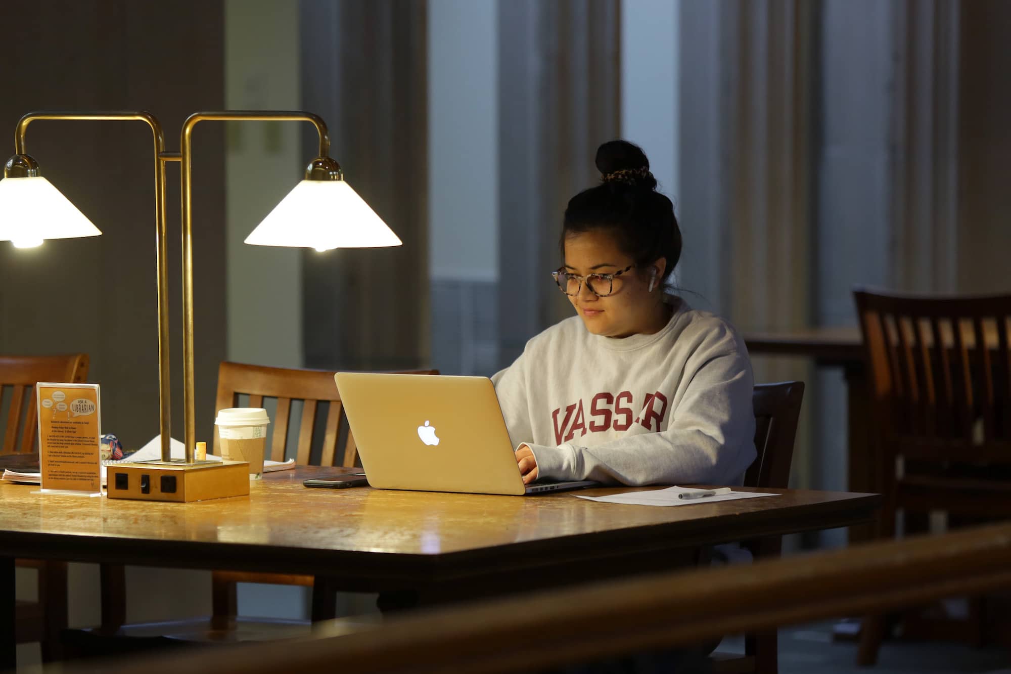 A student wearing a gray Vassar sweatshirt sits at a table and works on a laptop.